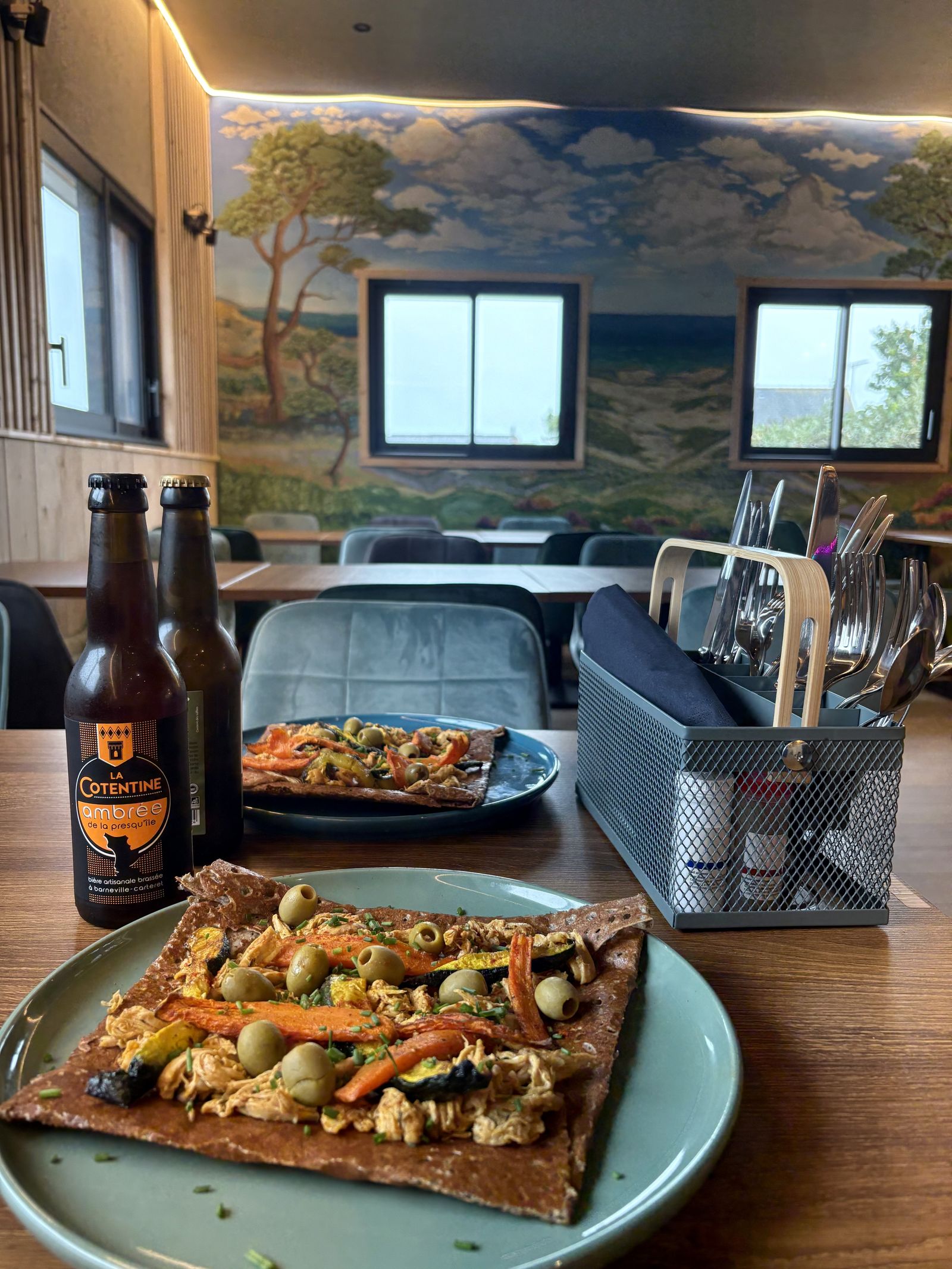 A galette served at the table with a bottle of Cotentine beer, the dune mural behind.