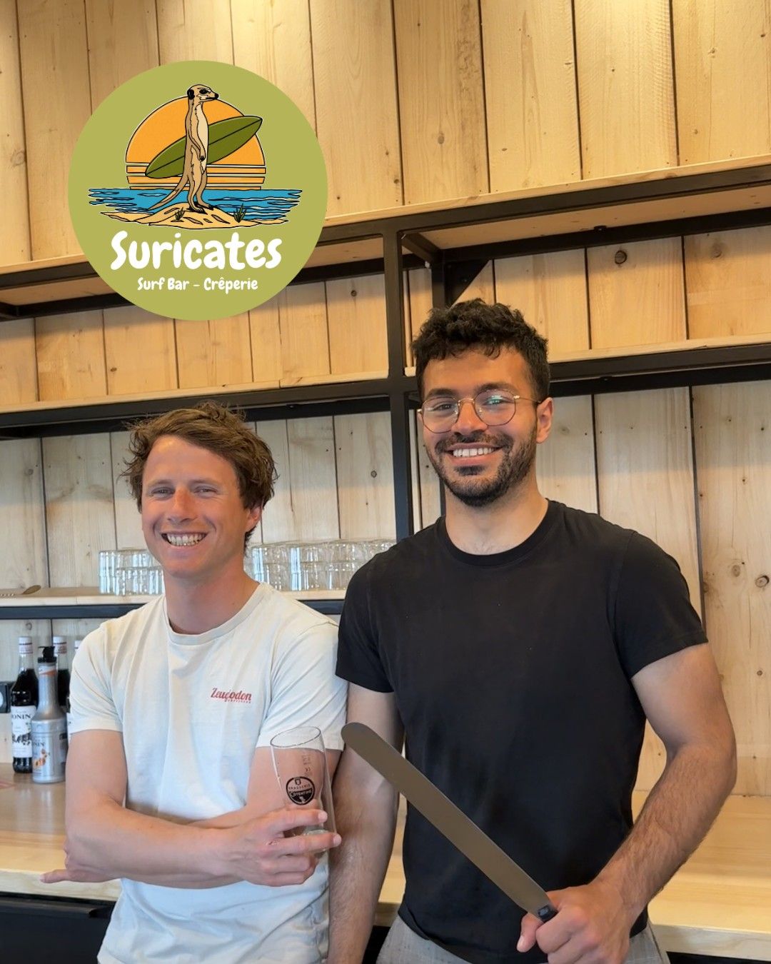 Henri and Zak, smiling, behind the wooden counter at Les Suricates, under the logo.