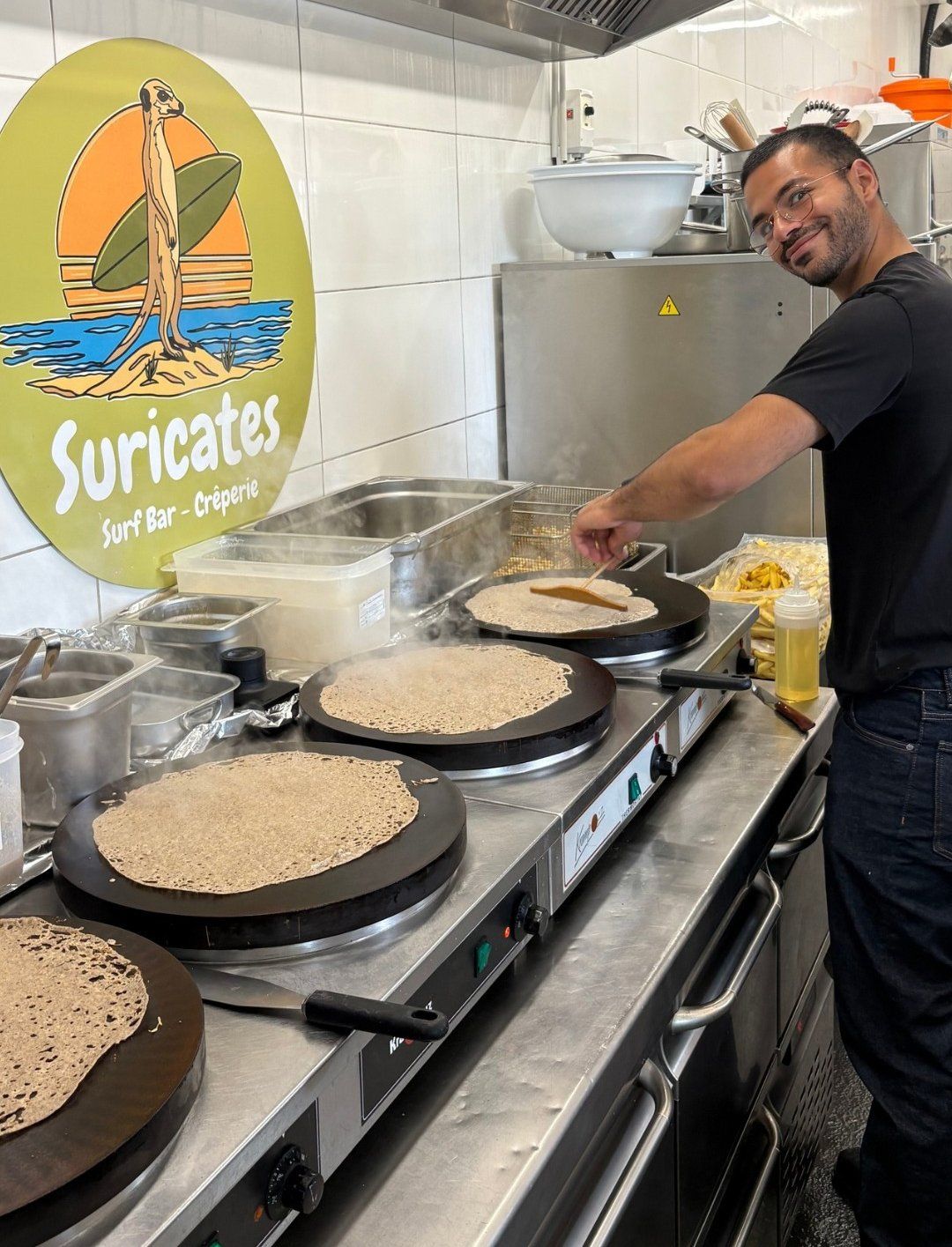 Zak in the kitchen, spatula in hand, in front of four biligs cooking buckwheat galettes.